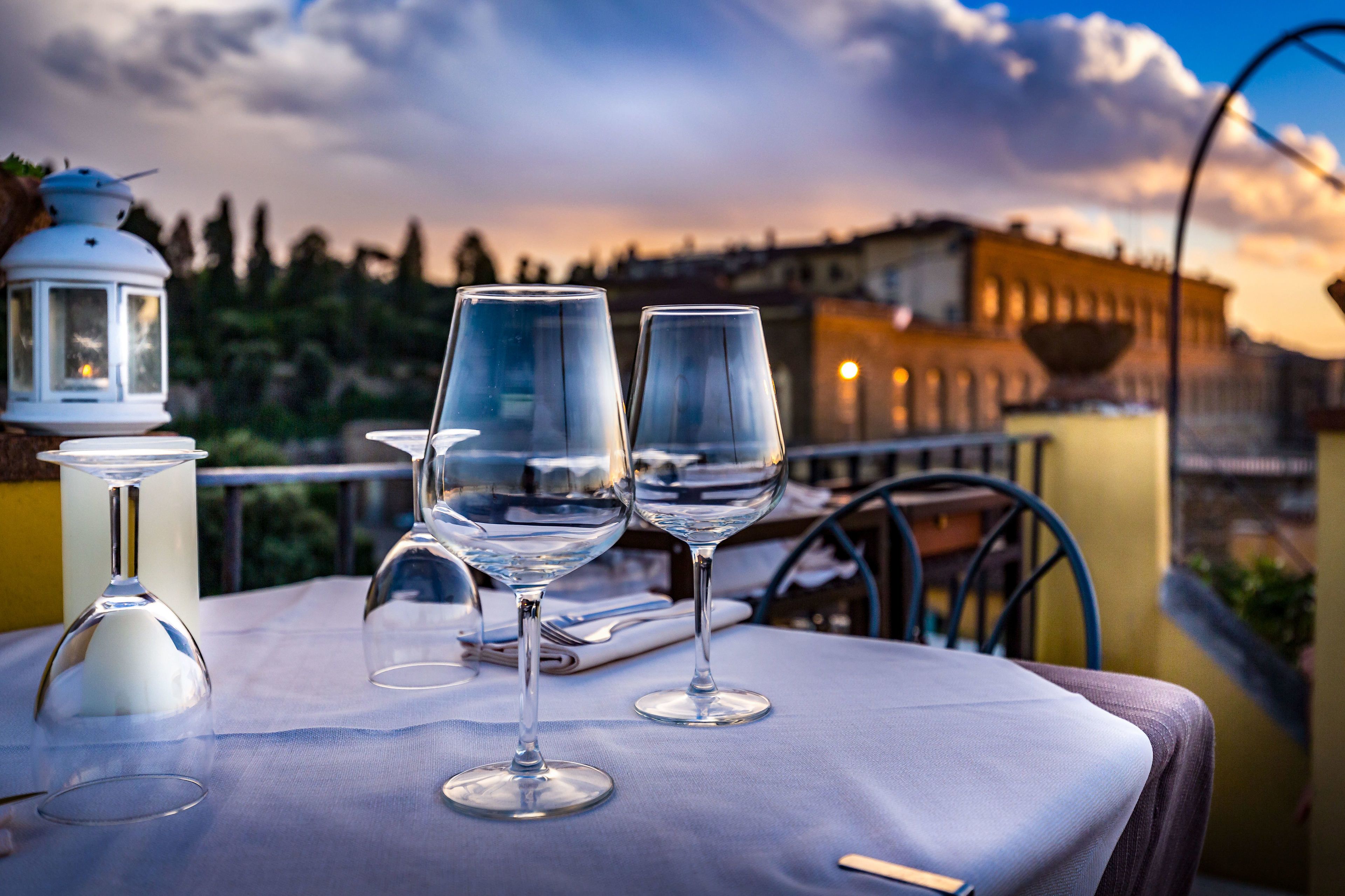 A table with two wine glasses and a candle on a balcony overlooking a cityscape at sunset.