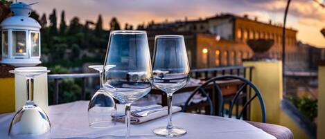 A table with two wine glasses and a candle on a balcony overlooking a cityscape at sunset.