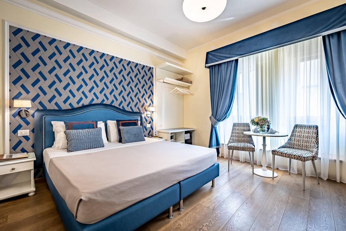 A hotel room with a large bed, a desk, a chair, and a small table with a vase of flowers.