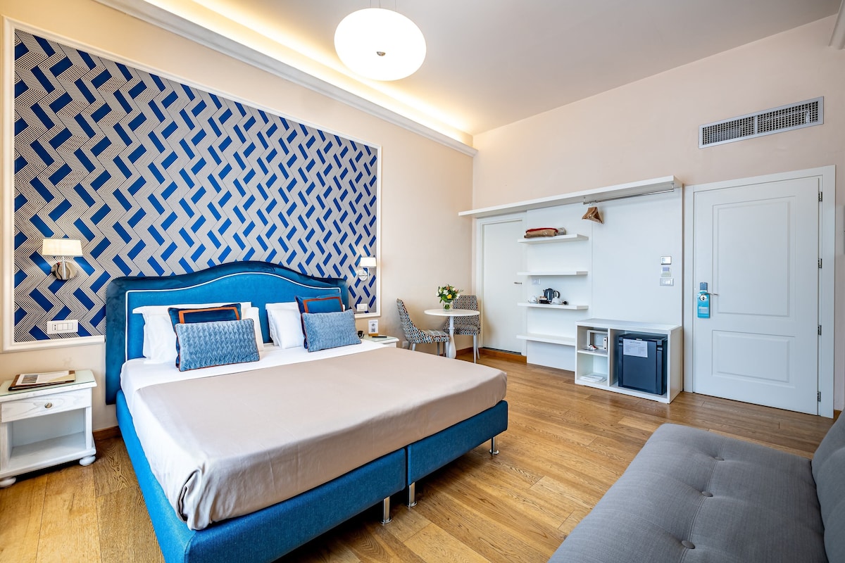 A modern hotel room with a large bed, a grey sofa, a small table, and a built-in wardrobe.