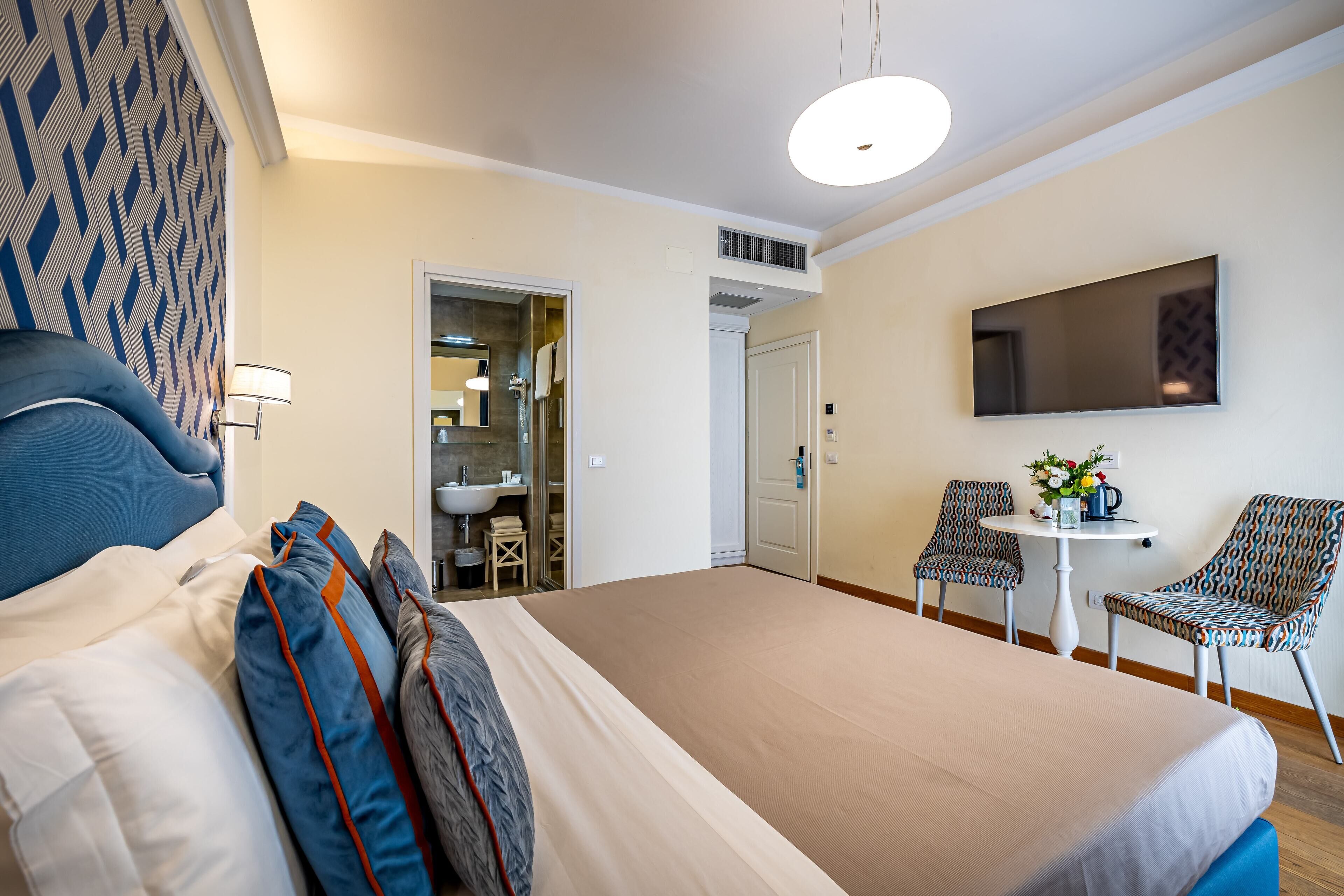 A hotel room with a large bed, a desk with two chairs, a TV, and a bathroom visible through an open door.