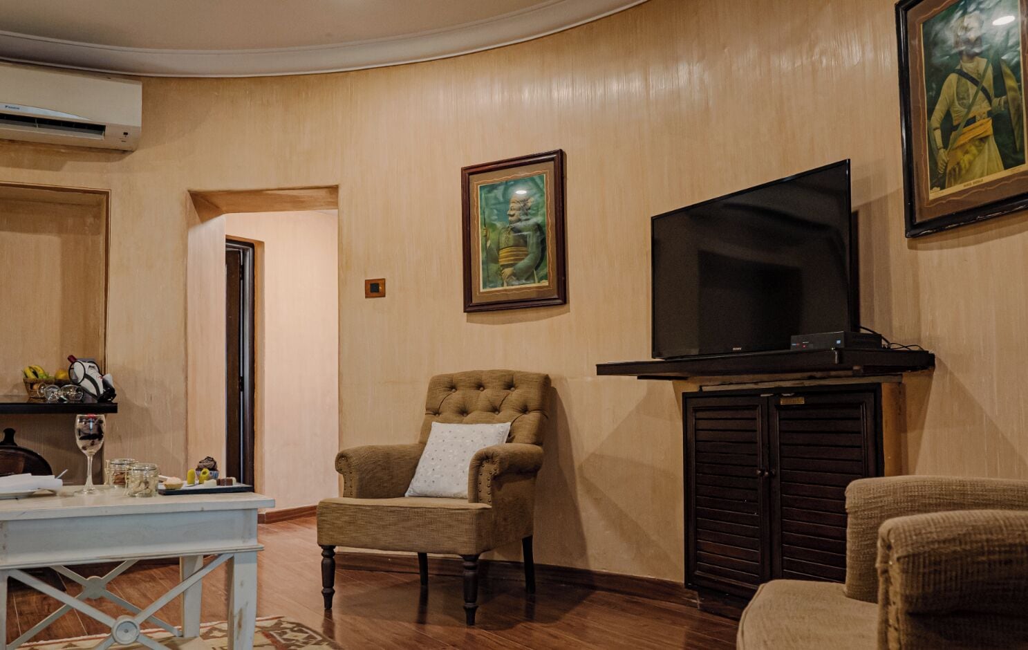 Una sala de estar con un televisor de pantalla plana grande, un sillón capitoné, una mesita con una copa de vino y un cuadro en la pared.