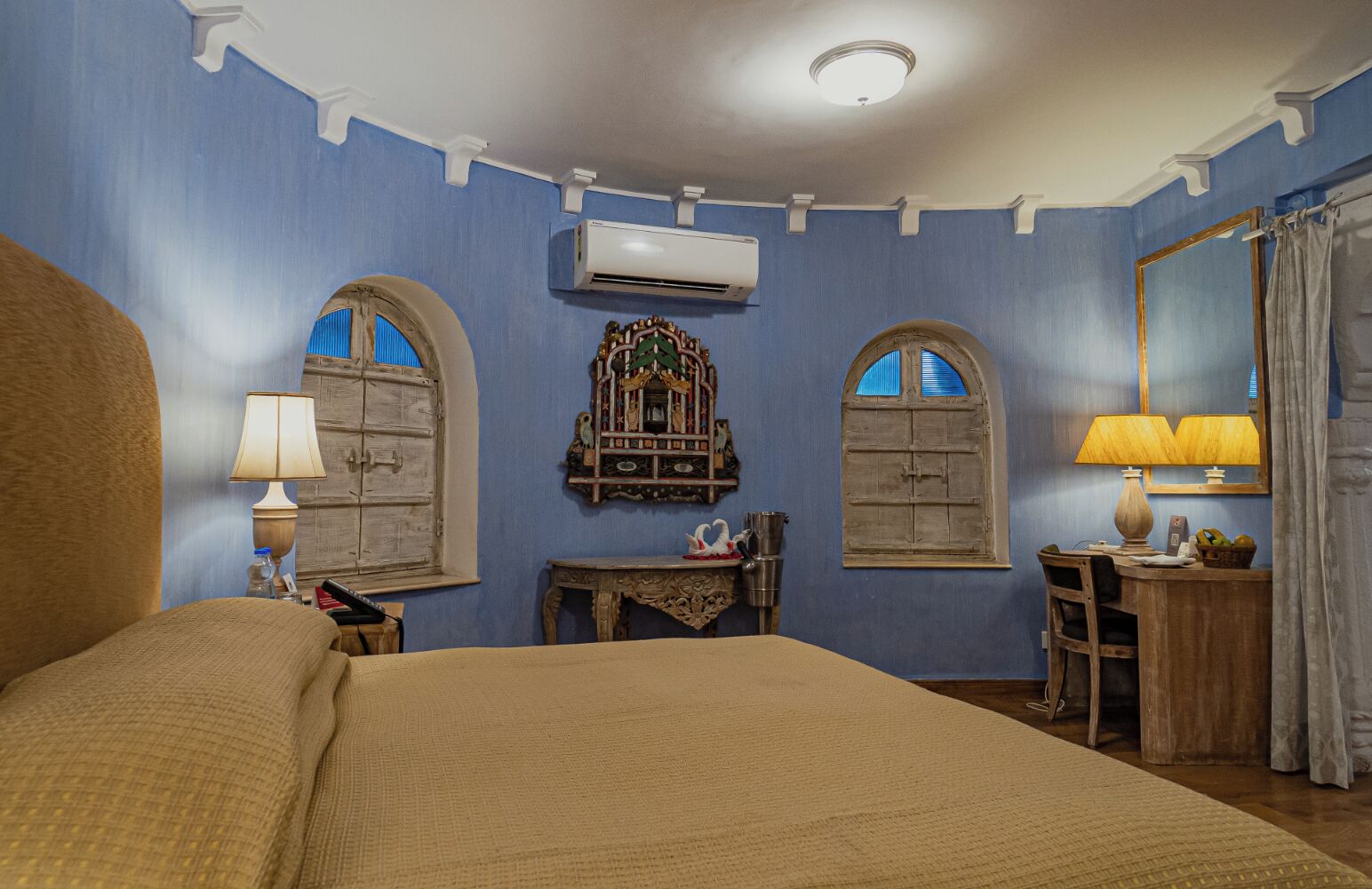 A room with a large bed, a desk with a lamp, an air conditioning unit, and a mirror.