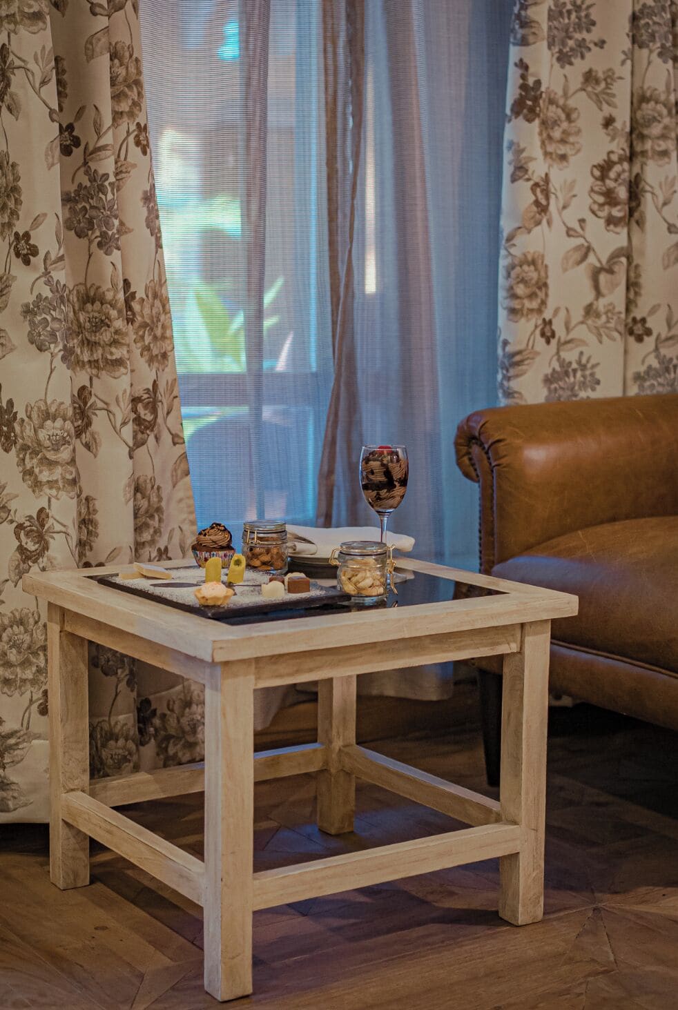 Una mesa de centro de madera con un telón de fondo estampado con motivos florales, un vaso de chocolate y un sillón de cuero marrón.
