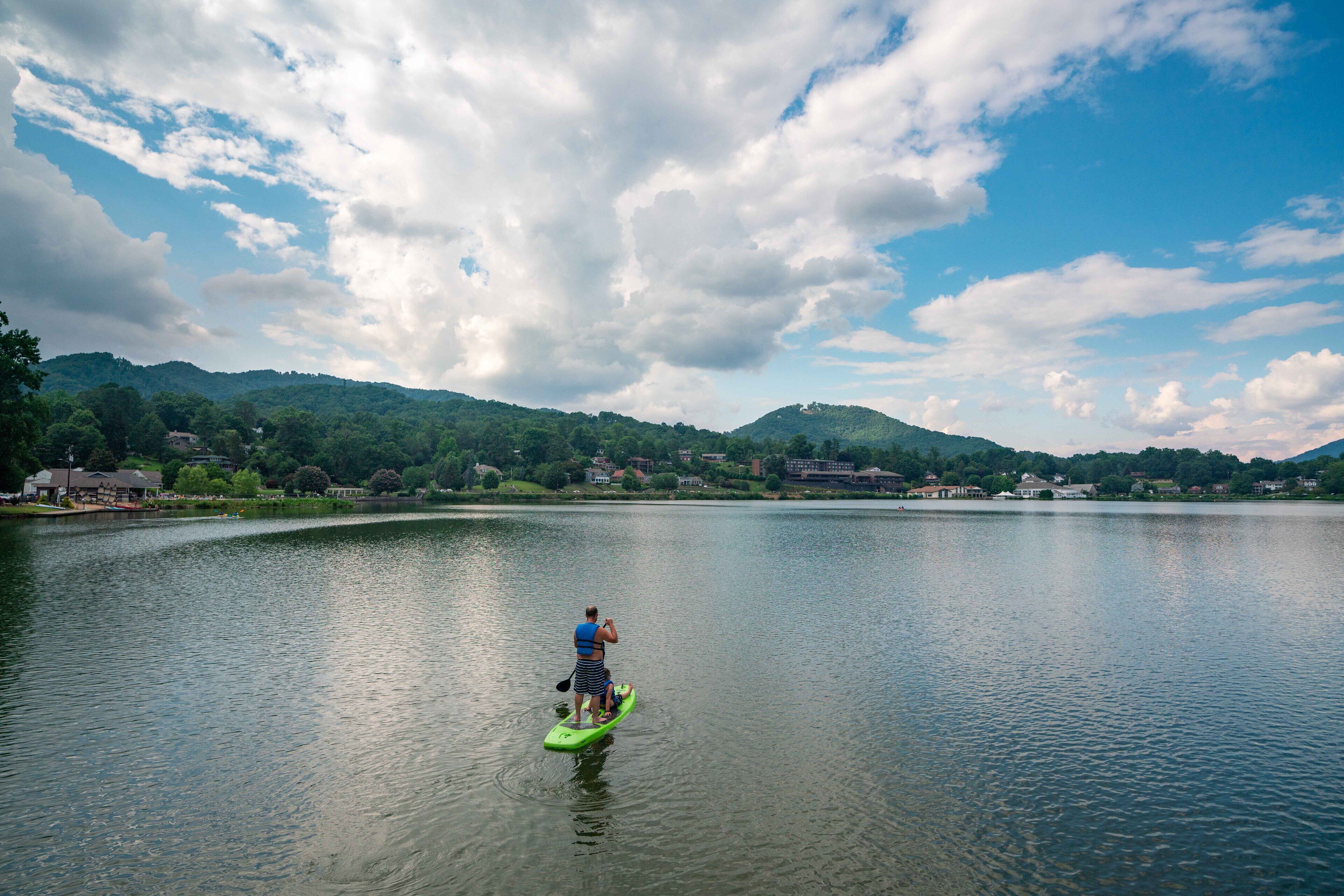 Una persona en una tabla de remo verde en un lago tranquilo, con casas y montañas al fondo.