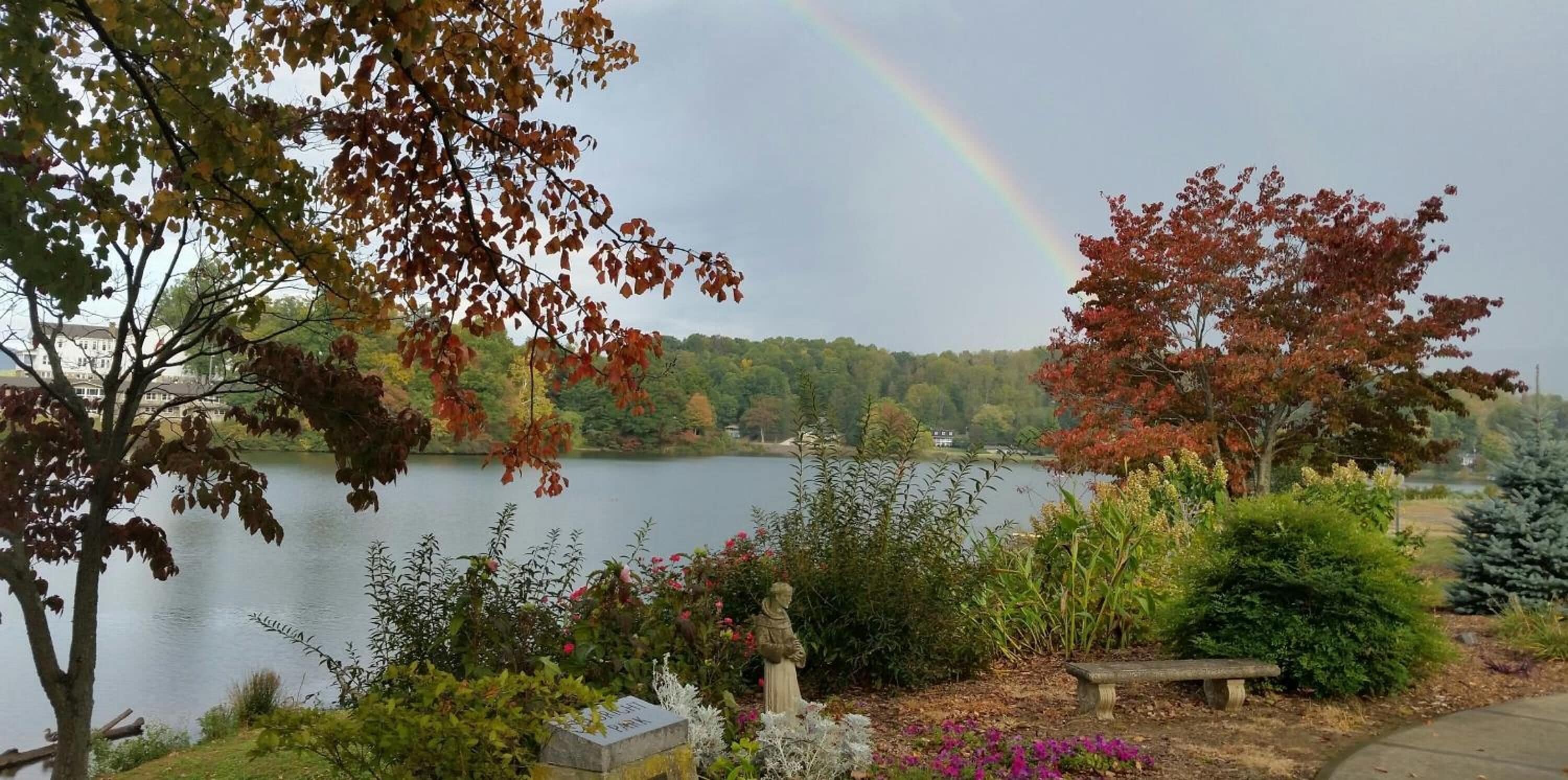 A lakeside view with autumn foliage, a rainbow, and a statue in the foreground.