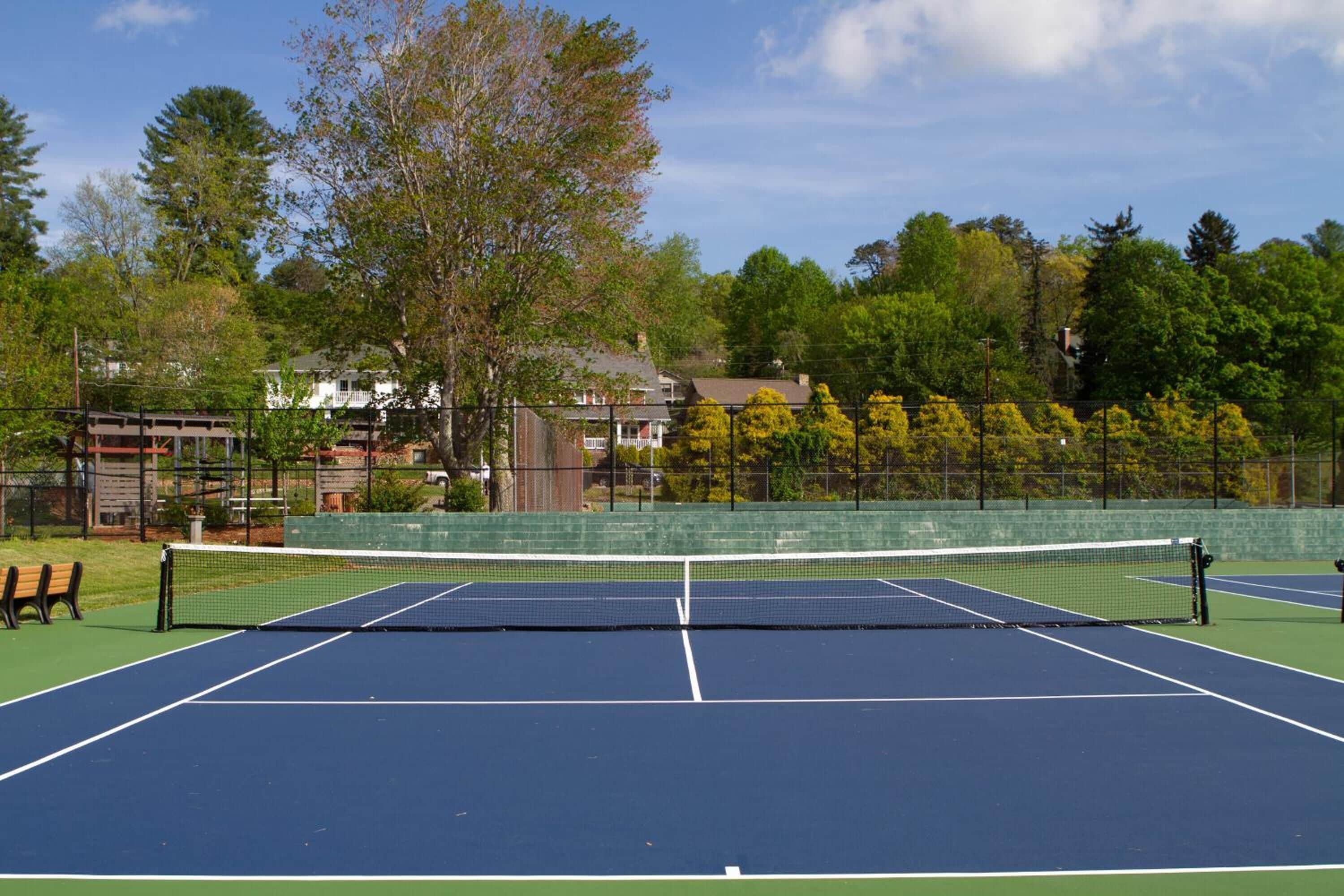 An outdoor tennis court with a net, surrounded by a fence and trees.