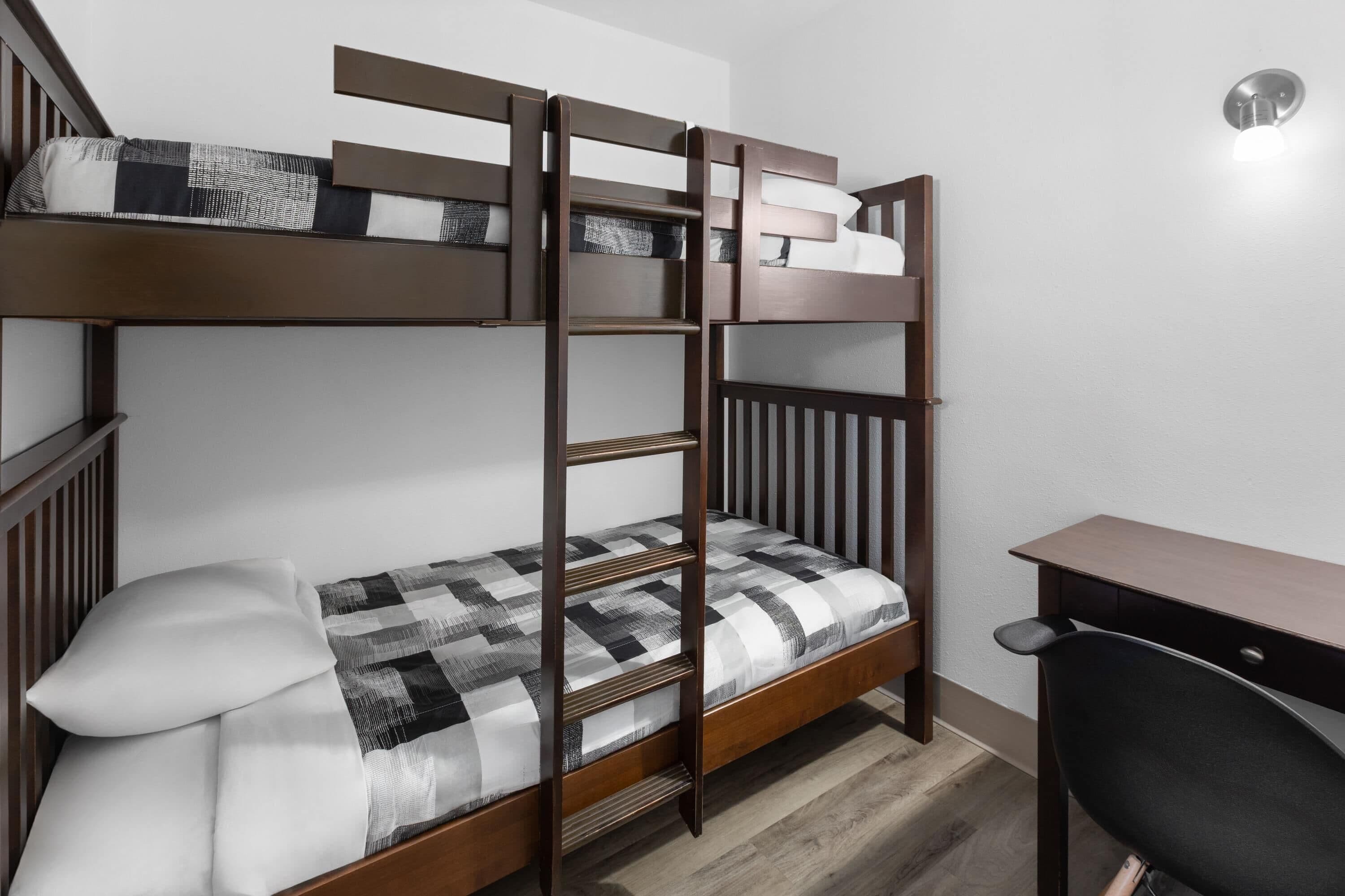 Una cama litera con escritorio y silla, un almohadón y una manta a cuadros.