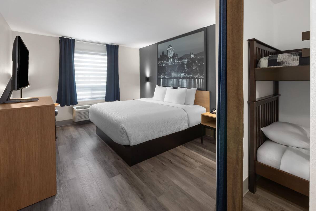 A modern hotel room with a large bed, a desk with a flat-screen TV, a cityscape artwork on the wall, and a bunk bed.