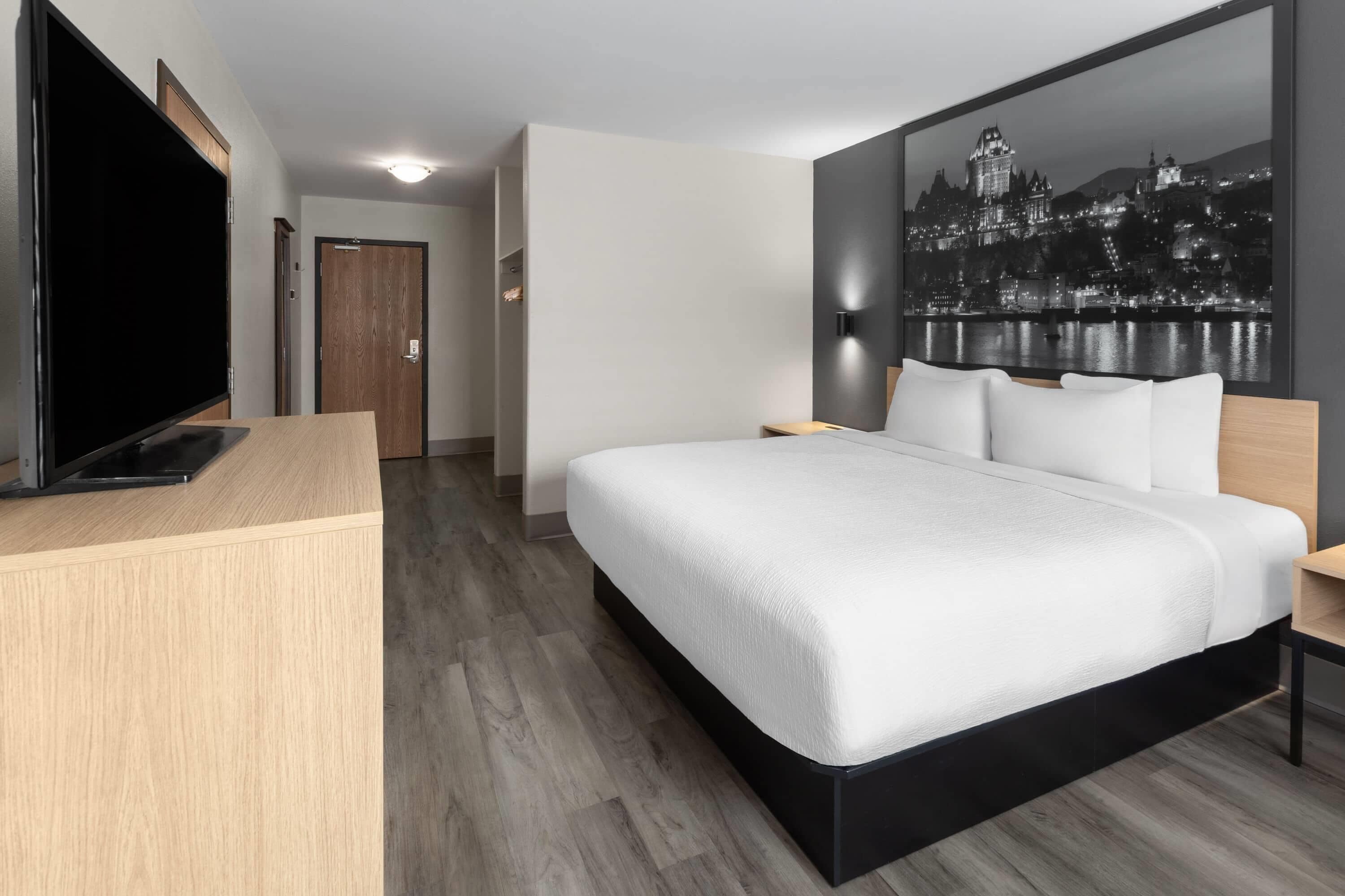 A modern hotel room with a large bed, a flat-screen TV, a wooden desk, and a cityscape painting on the wall.