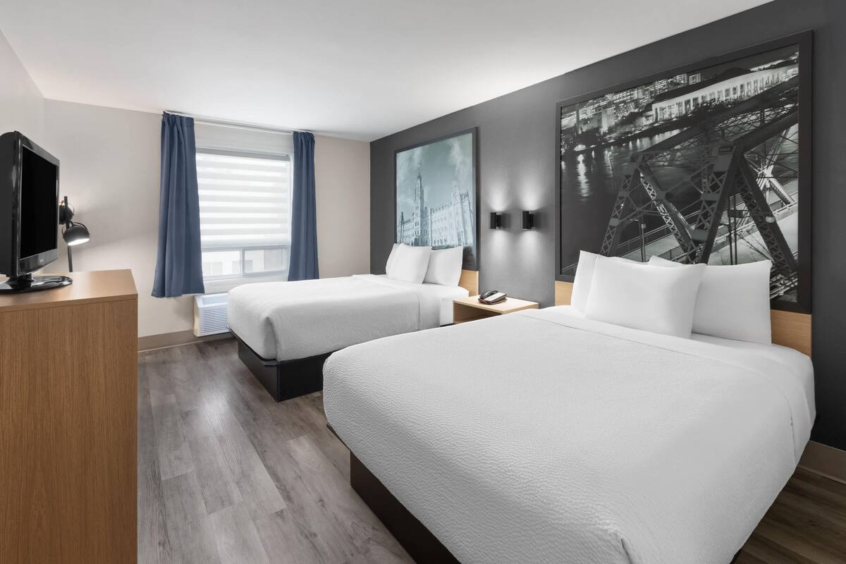 A hotel room with two beds, a desk, a TV, and a large cityscape painting on the wall.