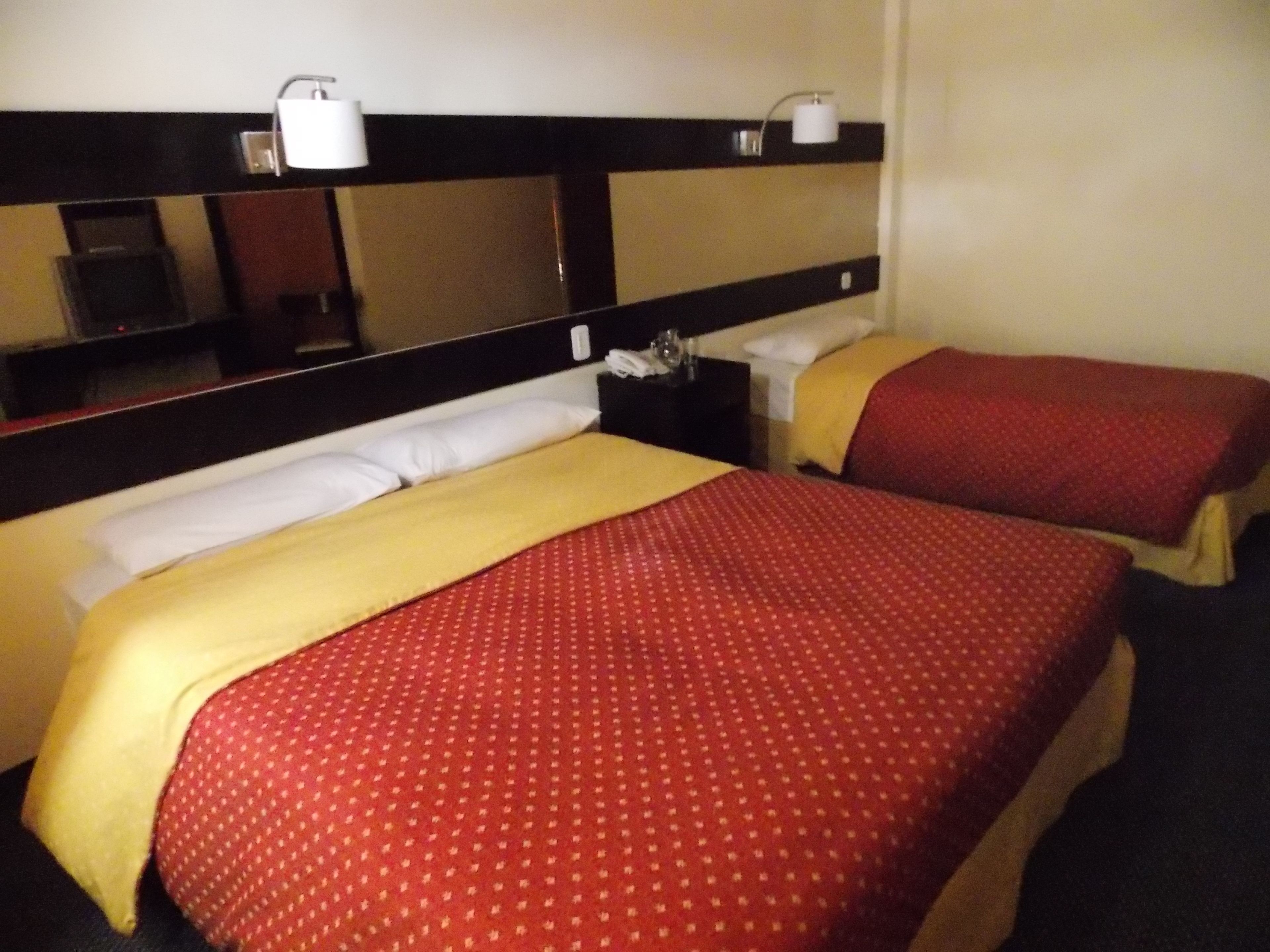Une chambre d’hôtel avec deux lits, chacun agrémenté de draps rouges et jaunes, un grand miroir au-dessus des lits et une petite table avec une lampe.