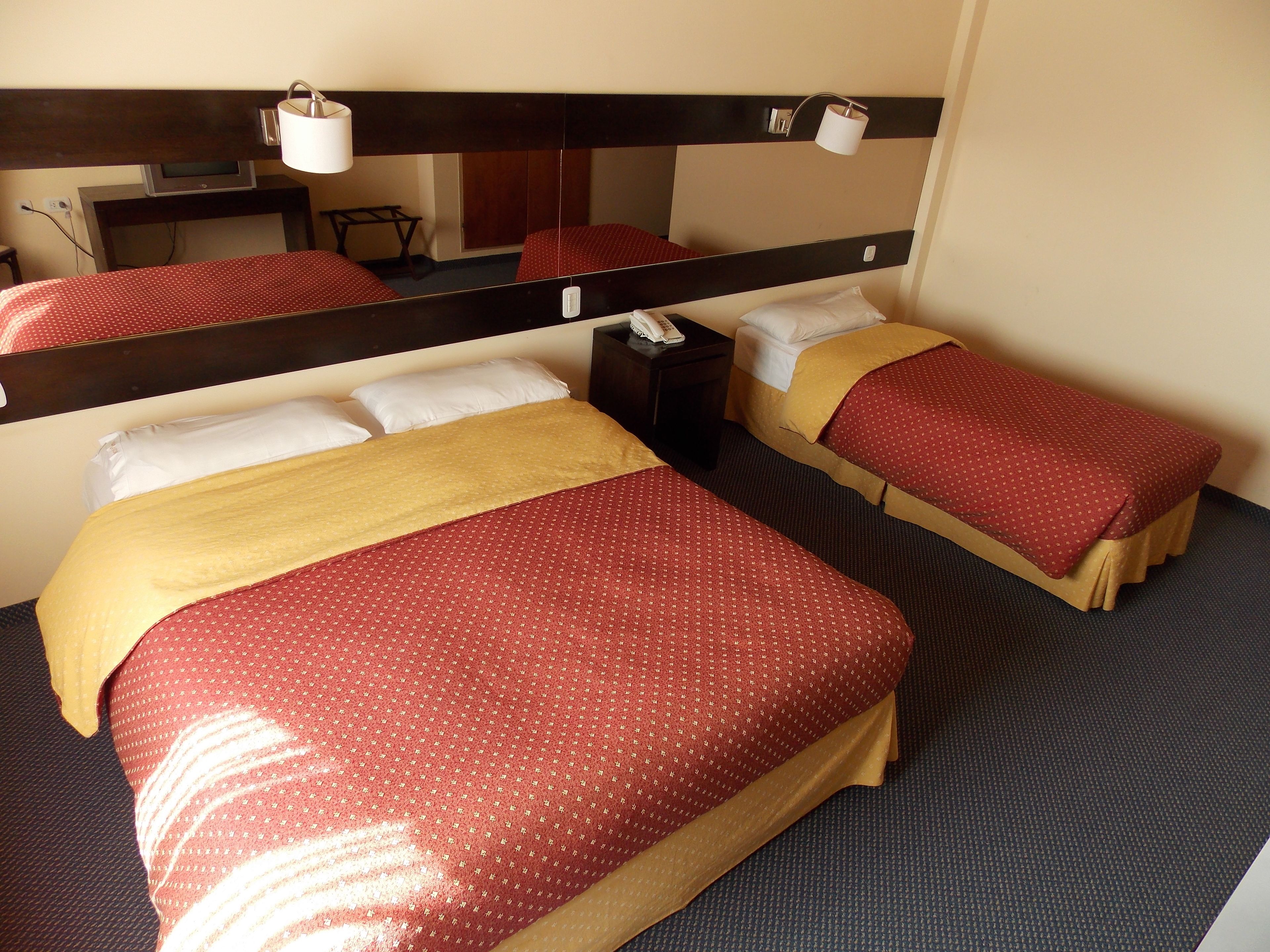 Habitación de hotel con dos camas, cada una con ropa de cama roja y amarilla, un espejo grande y una mesita de noche con lámpara.