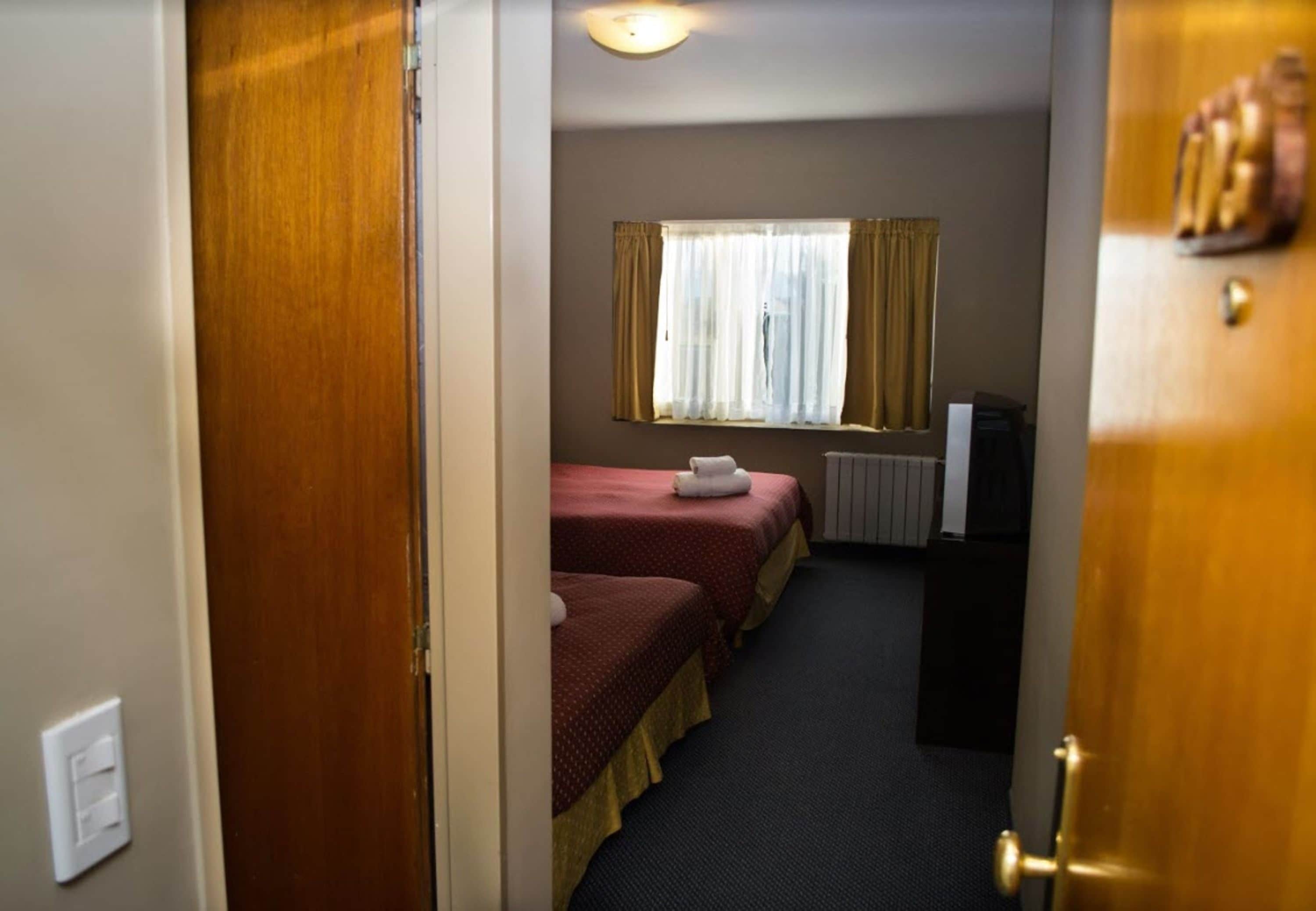 Une chambre d’hôtel étroite, avec deux lits, une fenêtre avec des rideaux, un radiateur et un téléviseur posé sur un meuble.