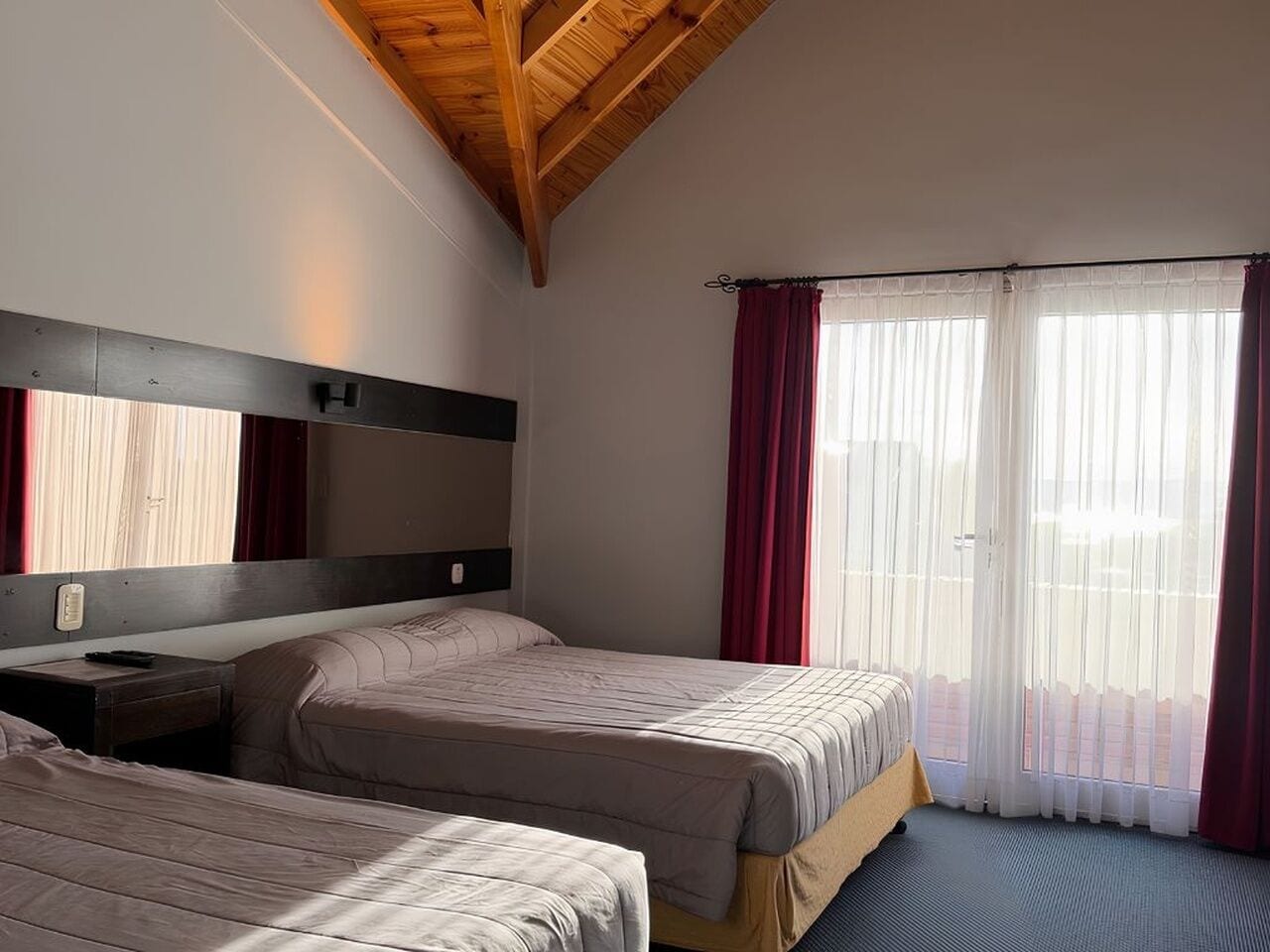 Habitación de hotel con dos camas, techo de madera, una ventana grande con cortinas y una mesita de noche.