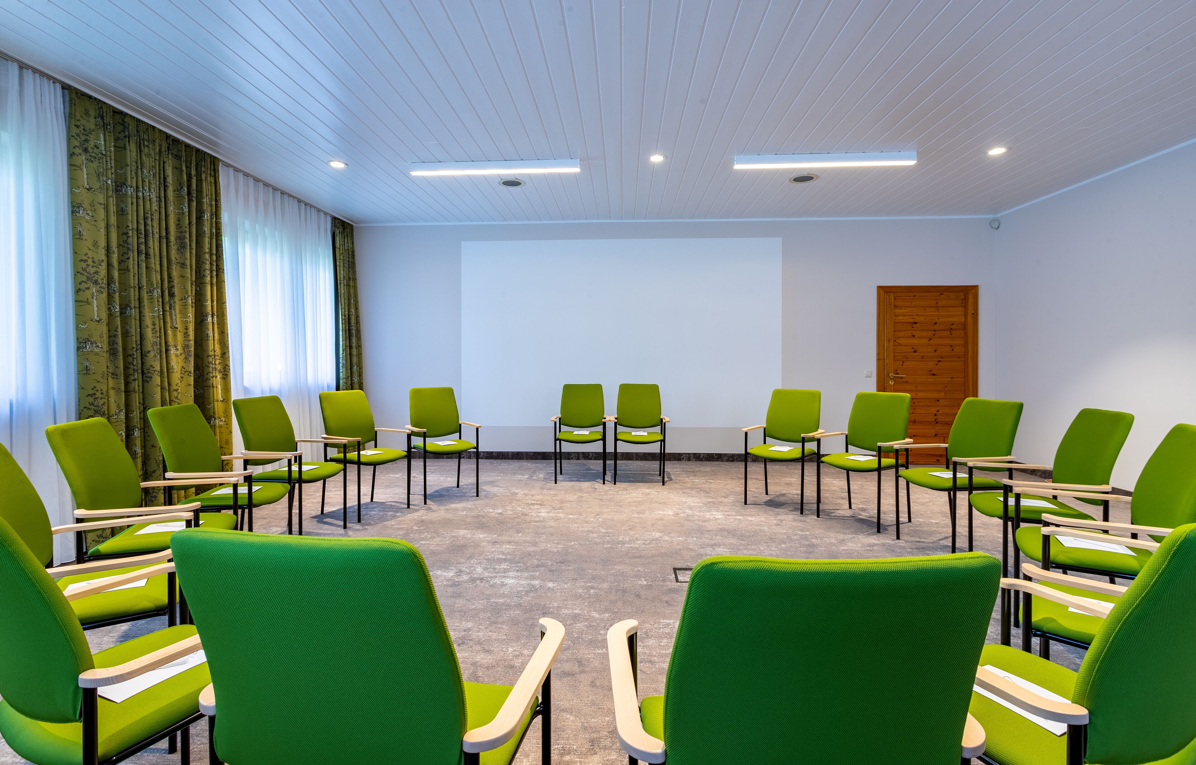 Ein Konferenzraum mit grünen, halbkreisförmig angeordneten Stühlen, weißen Wänden und einer Holztür.