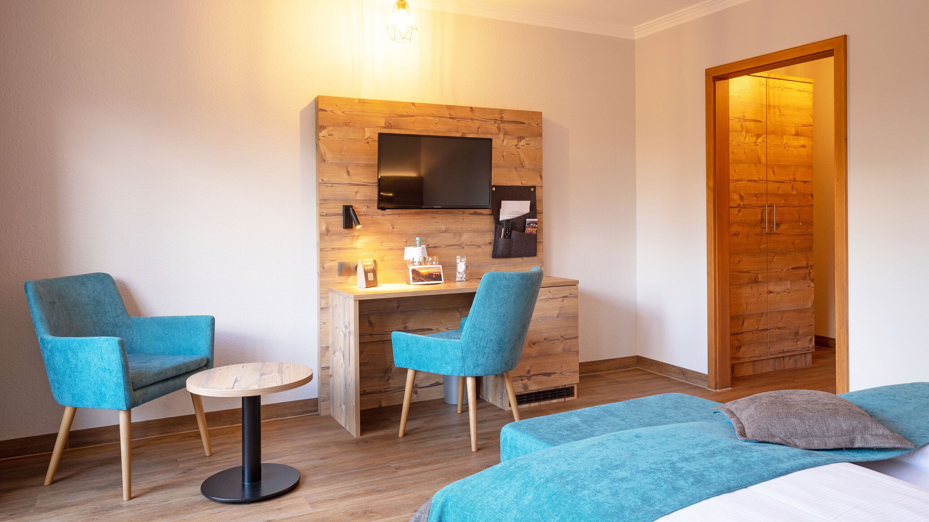 A modern hotel room with a wooden desk, two blue armchairs, a small round table, and a bed with a blue blanket.