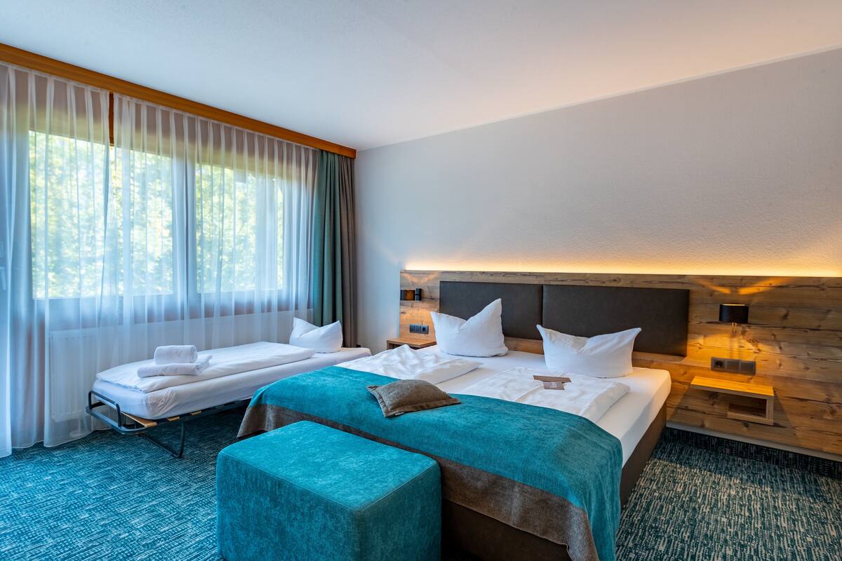Een hotelkamer met twee bedden, een groot raam met dunne gordijnen, een houten hoofdbord en een klein nachtkastje met een lamp.