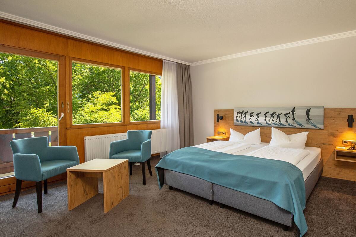 Een hotelkamer met een bed, een bureau, twee stoelen, een raam met uitzicht op bomen en een schilderij aan de muur.