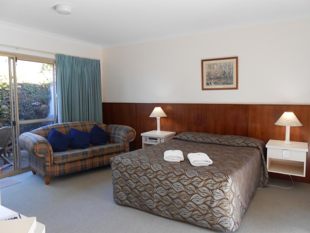A hotel room with a bed, a sofa, two bedside tables with lamps, a window with curtains, and a framed picture on the wall.