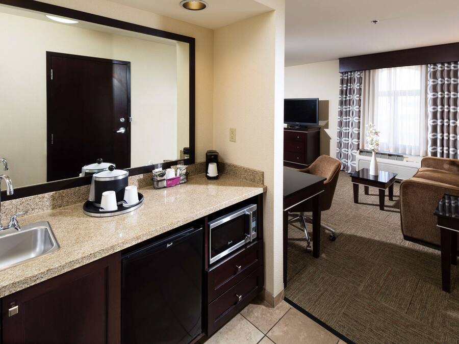 Quarto de hotel com cozinha compacta, um espelho grande, área de jantar com mesa e cadeiras, televisão e janela com cortinas.