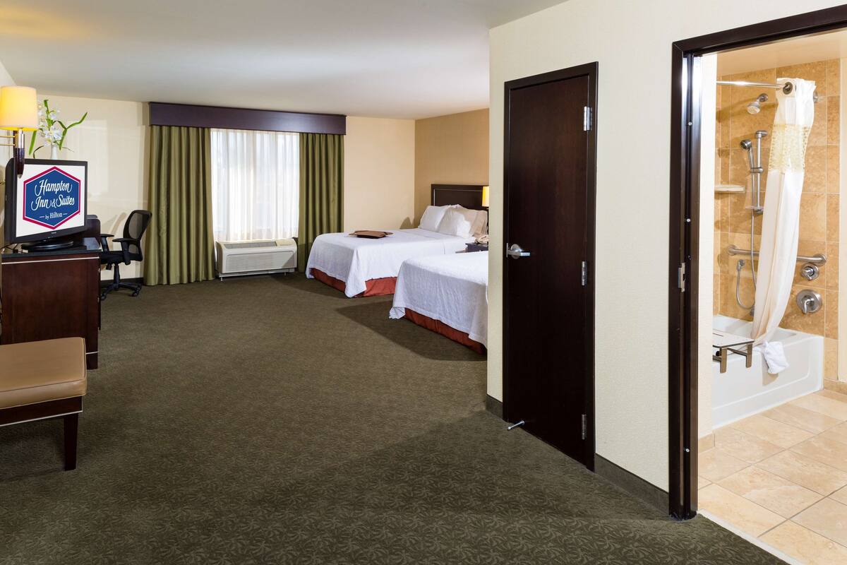 Quarto de hotel com duas camas, uma secretária com computador, uma cadeira, luminária de mesa, janela com cortinas e banheiro com banheira e chuveiro.