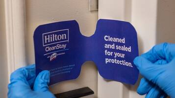 A person wearing blue gloves placing a blue sign on a door that reads "Hilton CleanStay" and "Cleaned and sealed for your protection."