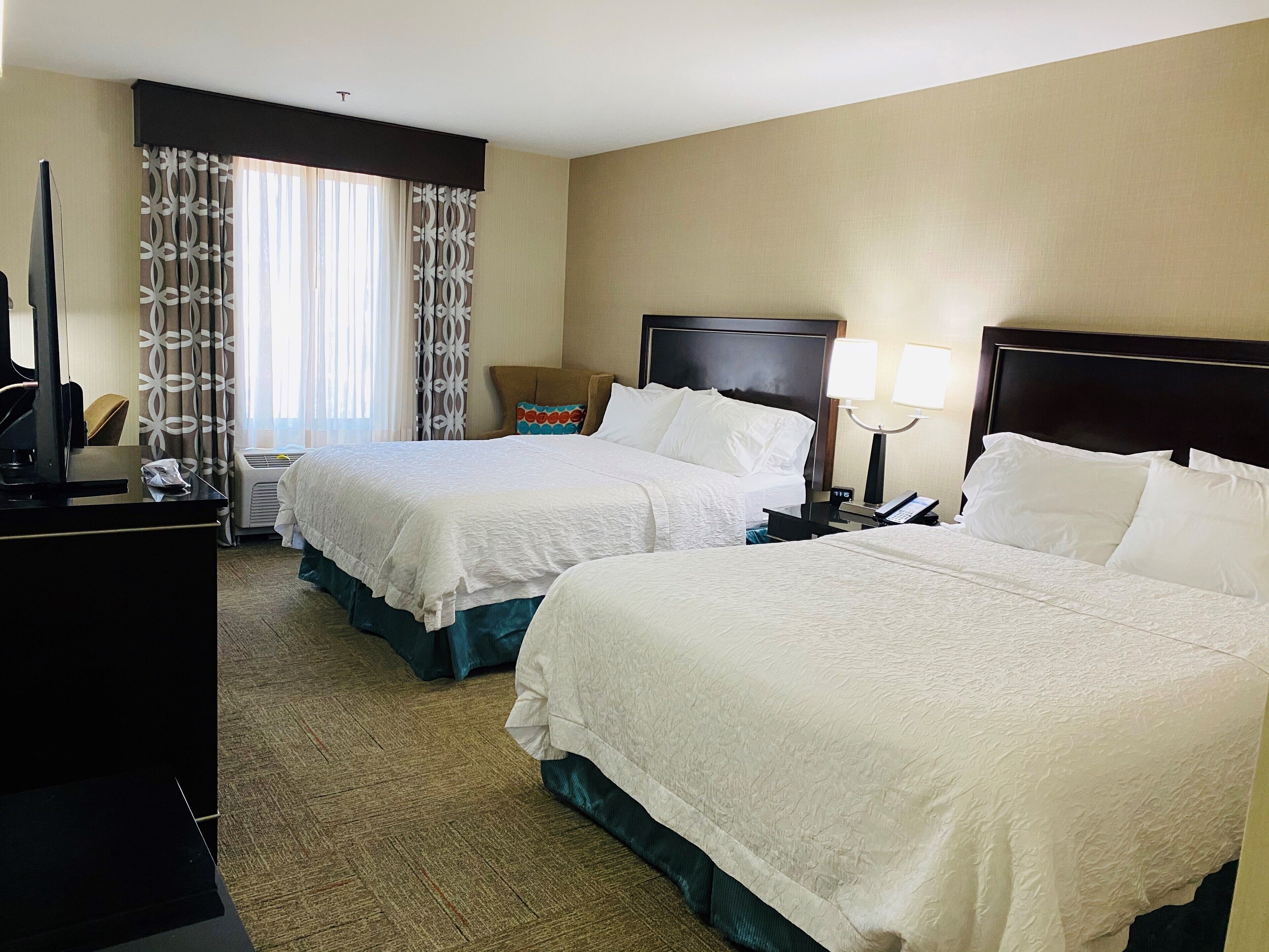 A hotel room with two beds, a desk, a chair, a TV, and a window with curtains.