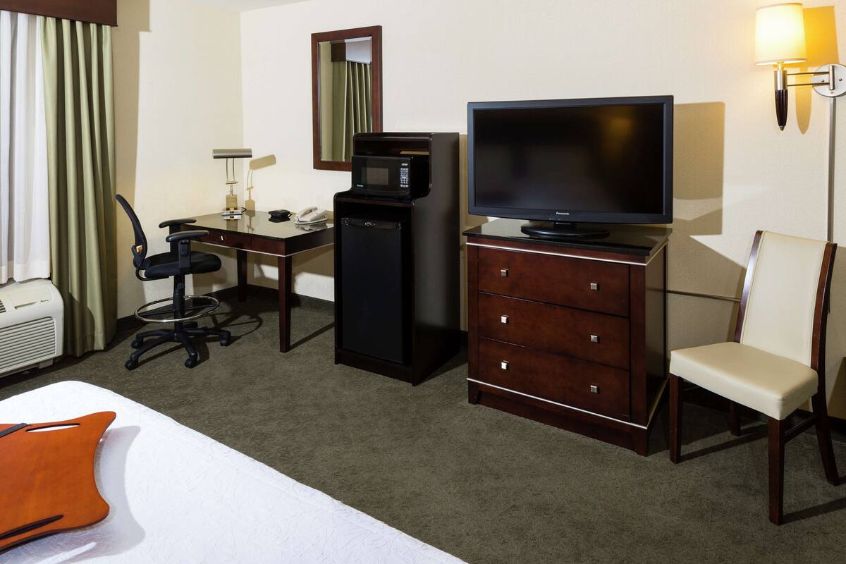 Quarto de hotel com cama, escrivaninha com computador, televisão, micro-ondas, cadeira e abajur.