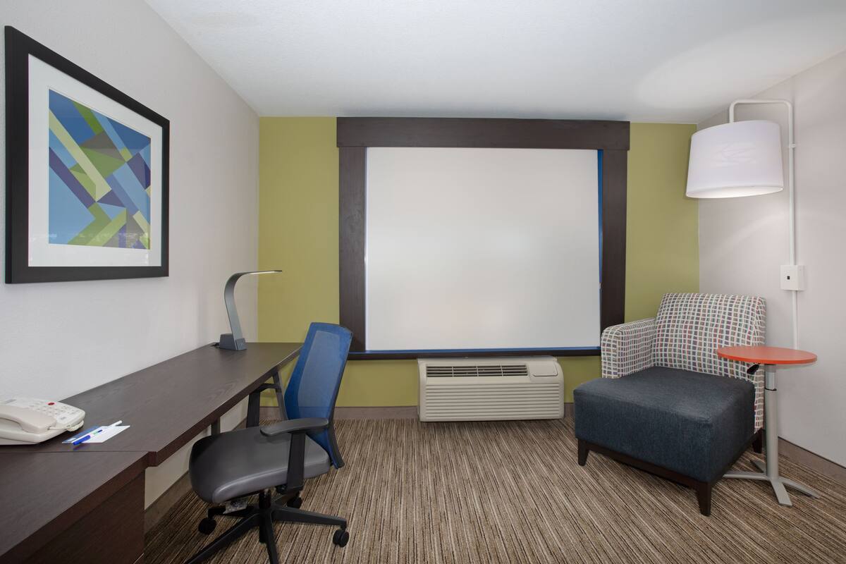 A hotel room with a desk, chair, ottoman, and a small table with a lamp.