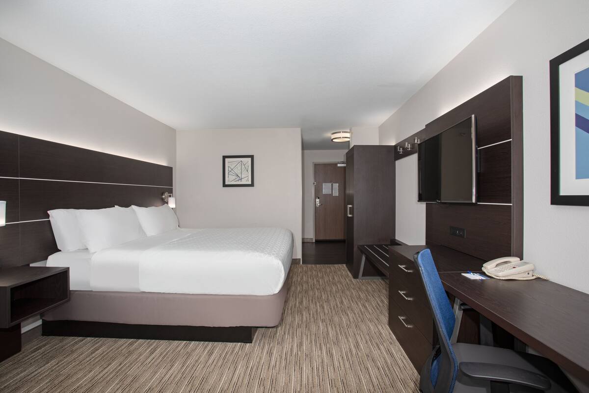 A modern hotel room with a large bed, a desk with a telephone, a chair, and a wardrobe.