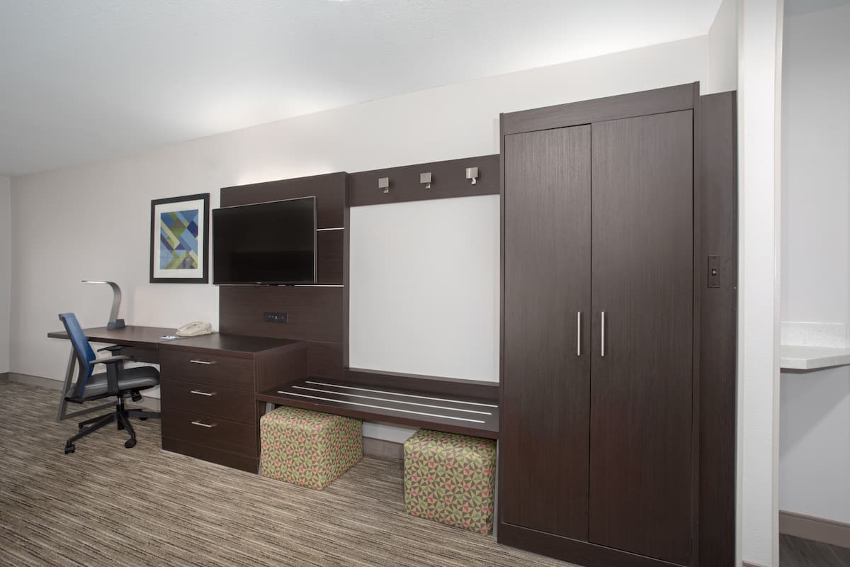 A hotel room with a dark wooden desk, a flat-screen TV, a large wardrobe, and patterned ottomans.