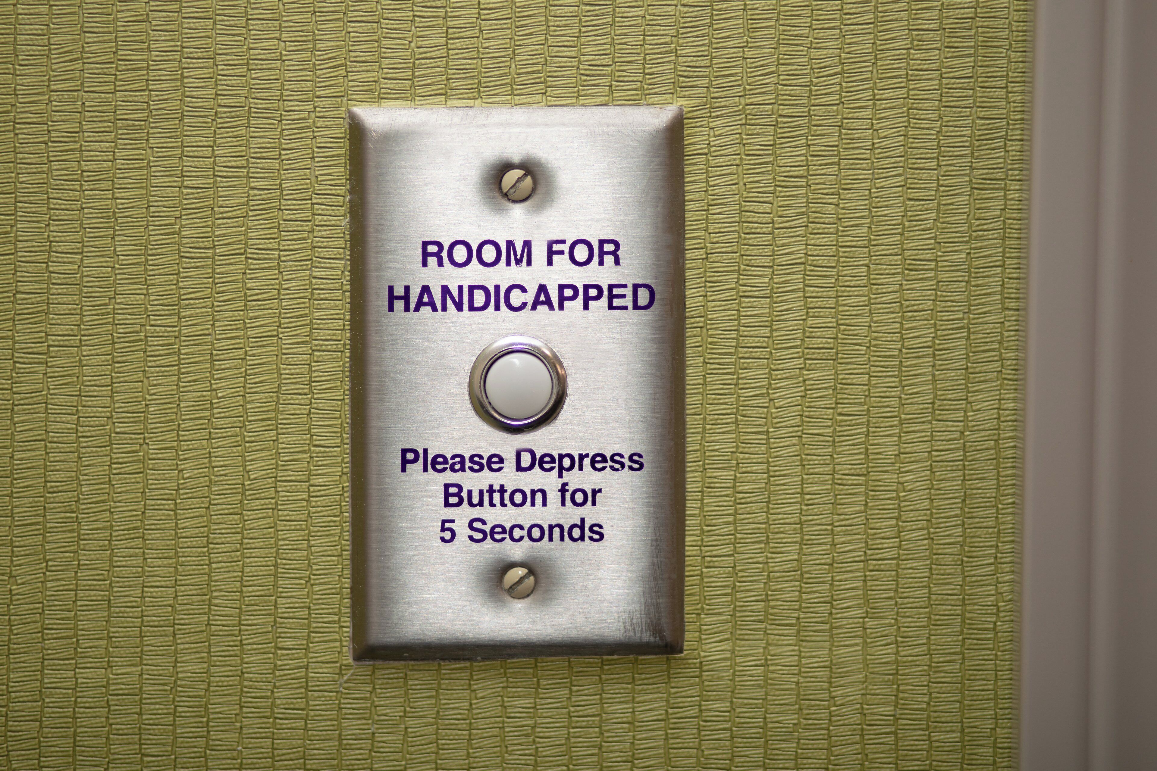 Una señal metálica con la inscripción 'ROOM FOR HANDICAPPED' e instrucciones para presionar un botón.