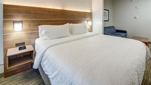 A hotel room with a large bed, a wooden headboard, a bedside table with a clock, and a small seating area with a chair and a table. - Holiday Inn Express Hotel & Suites Quincy I-10 by IHG (Quincy)