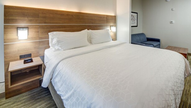 A hotel room with a large bed, a wooden headboard, a bedside table with a clock, and a small seating area with a chair and a table. - Holiday Inn Express Hotel & Suites Quincy I-10 by IHG (Quincy)