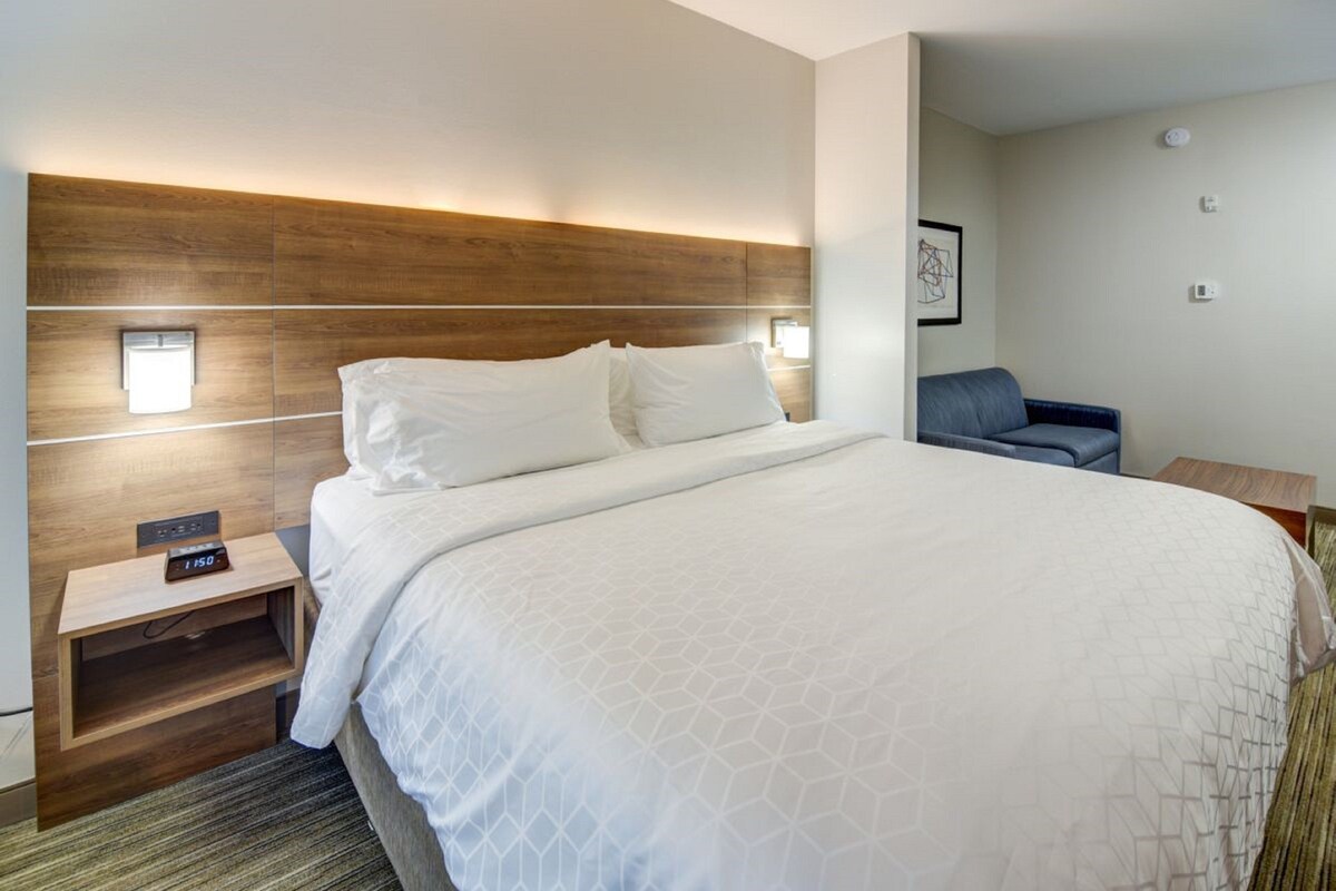 Una habitación de hotel con una cama grande, cabecera de madera, lámparas de noche, una mesita y una silla azul.