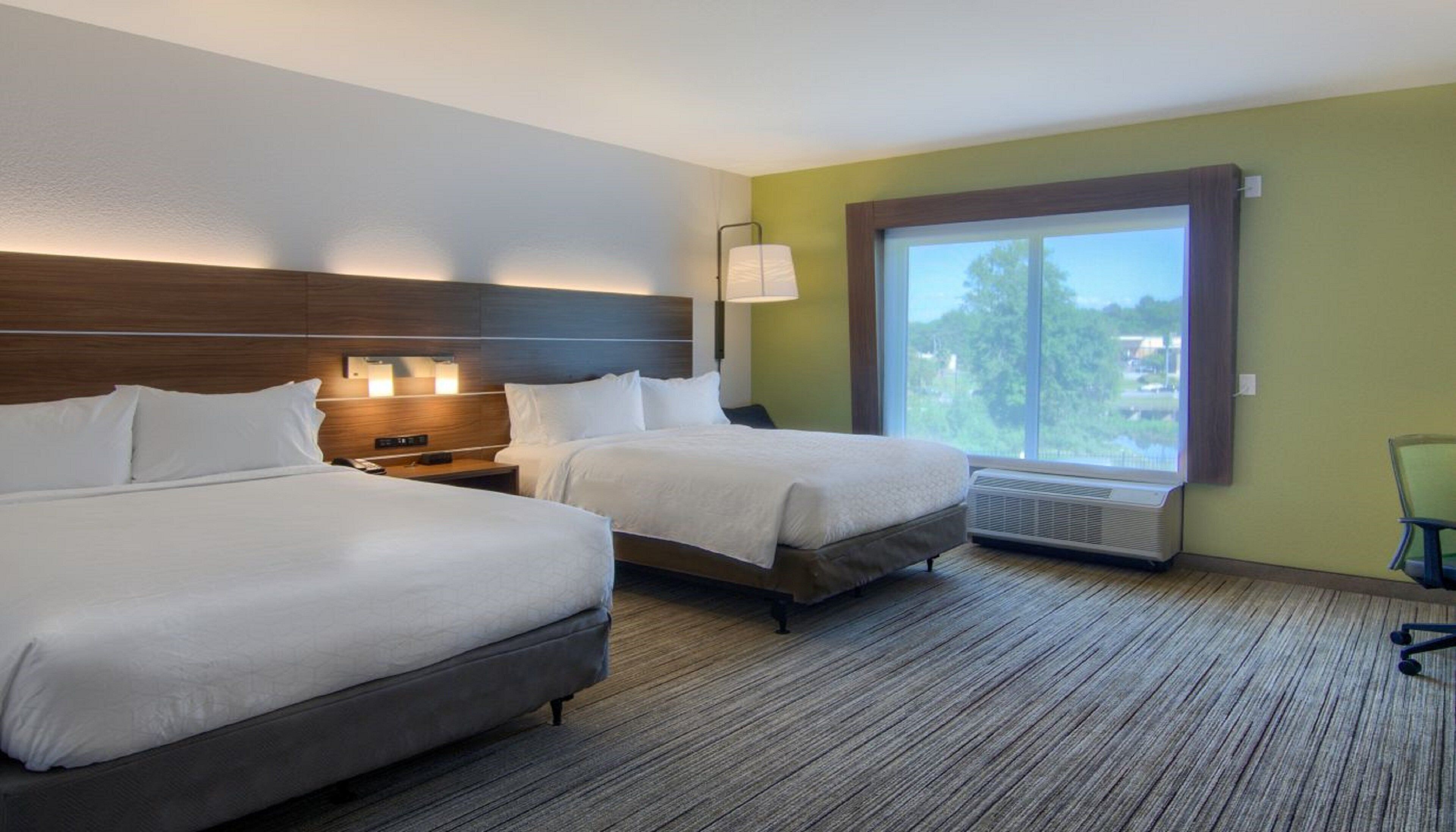 Habitación de hotel con dos camas, una mesita de noche con lámpara, un ventanal con vistas y una silla.