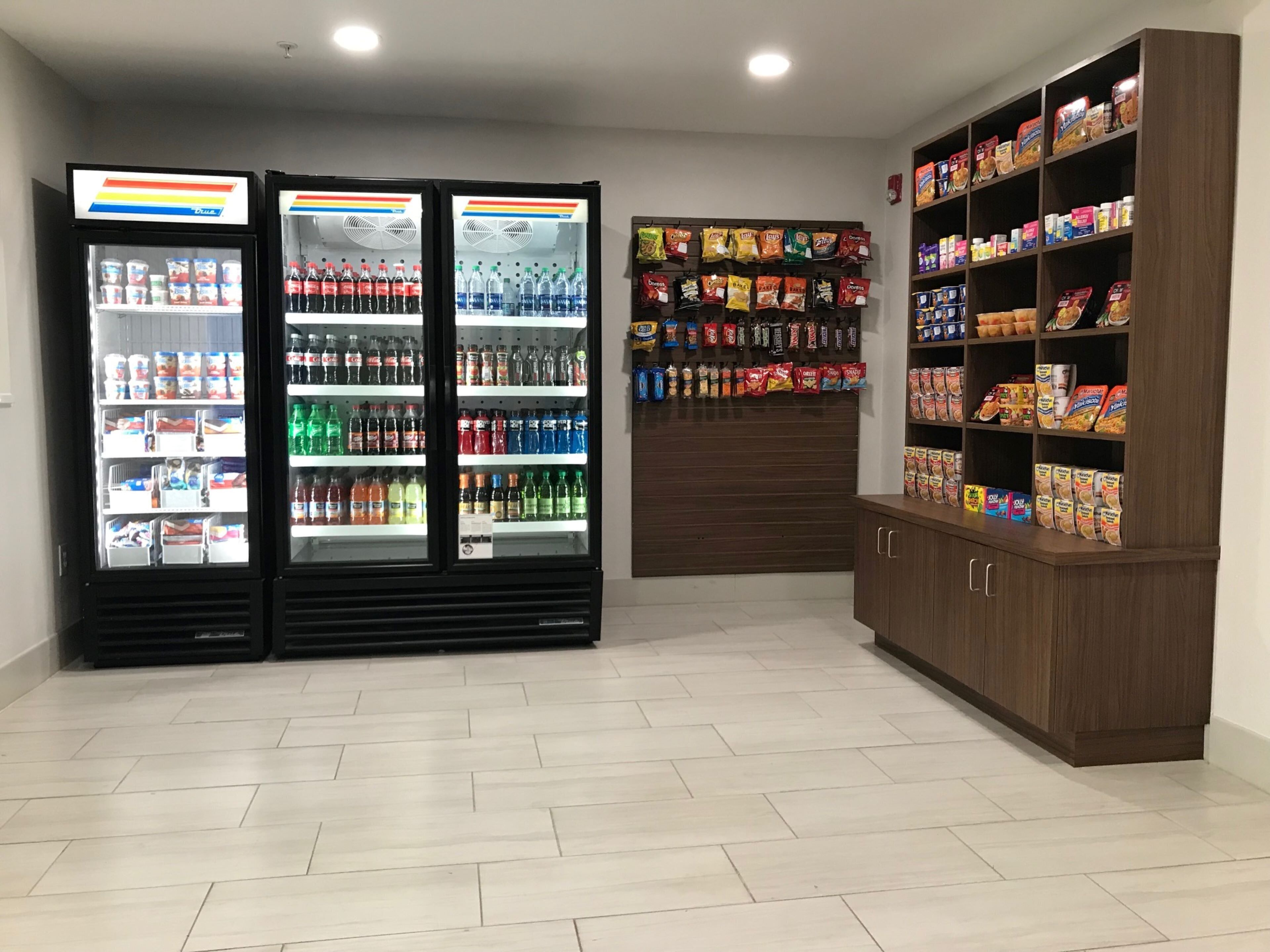 Una habitación con dos refrigeradores para bebidas llenos de varias opciones, una pared con aperitivos en bolsas colgantes y un armario con cajas de cereal.