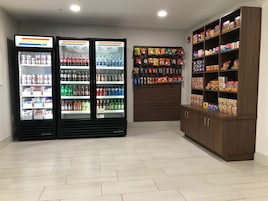 A room with two beverage refrigerators stocked with various drinks, a snack wall with hanging bags, and a cabinet with cereal boxes.