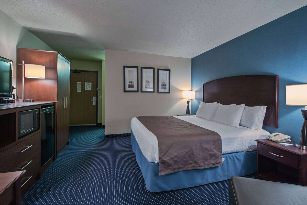 A hotel room with a bed, bedside table, lamp, and a desk with a lamp. There is a television on a cabinet and a door leading to another room.