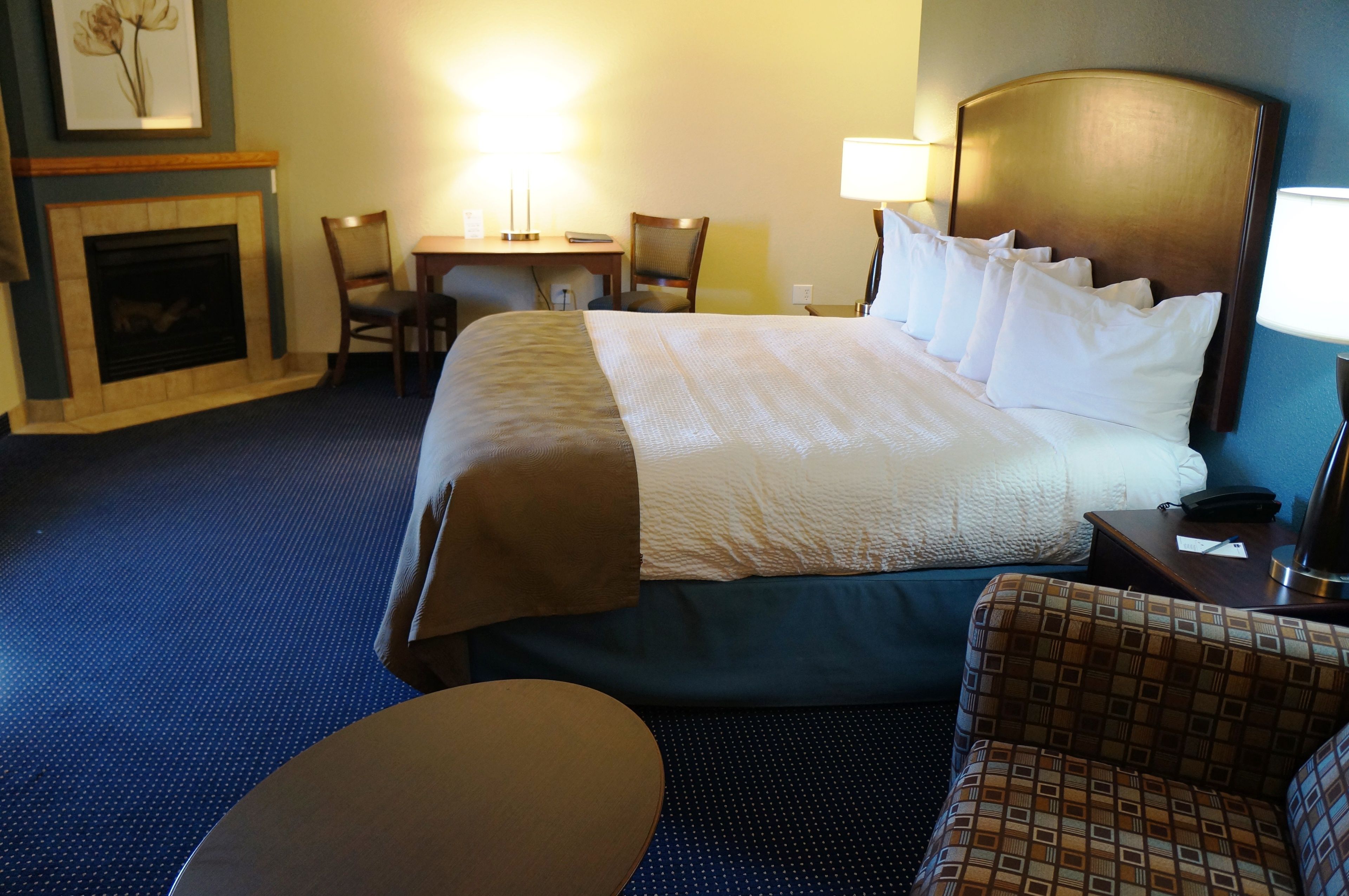 Quarto de hotel com uma cama grande, uma escrivaninha com cadeira, uma mesinha redonda e uma poltrona estampada.