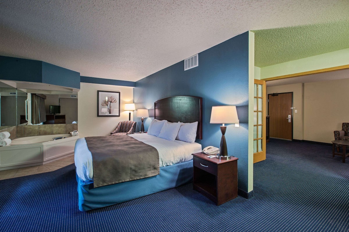 A hotel room with a large bed, bedside lamps, a nightstand, a desk with a lamp, a chair, a bathroom with a bathtub, and a mirror.