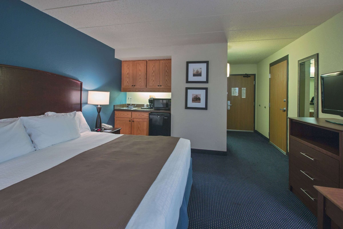 A hotel room with a large bed, a desk with a lamp, a kitchenette, and a television.