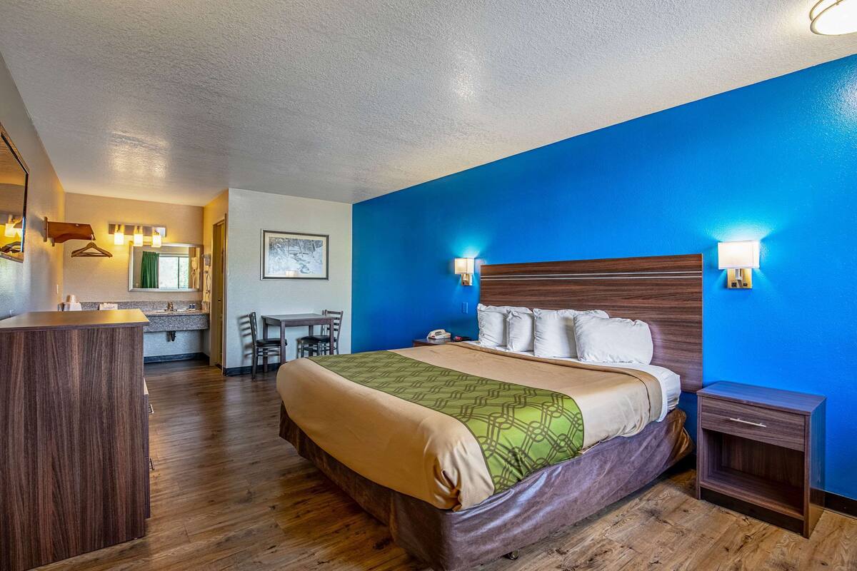 A hotel room with a wooden floor, a bed with a green and beige bedspread, a wooden headboard, two bedside lamps, a small wooden nightstand, and a bathroom visible in the background.