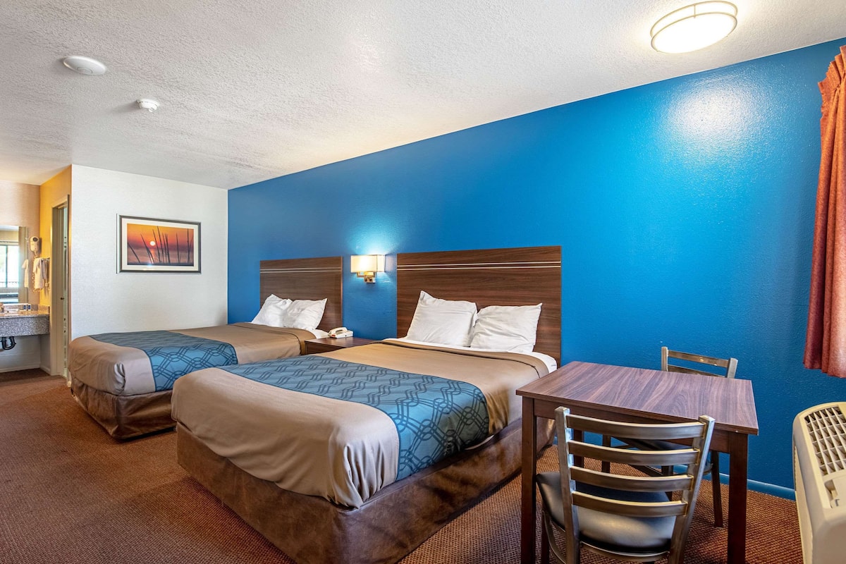 A hotel room with two beds, a wooden table, a chair, and a bathroom with a sink and mirror.