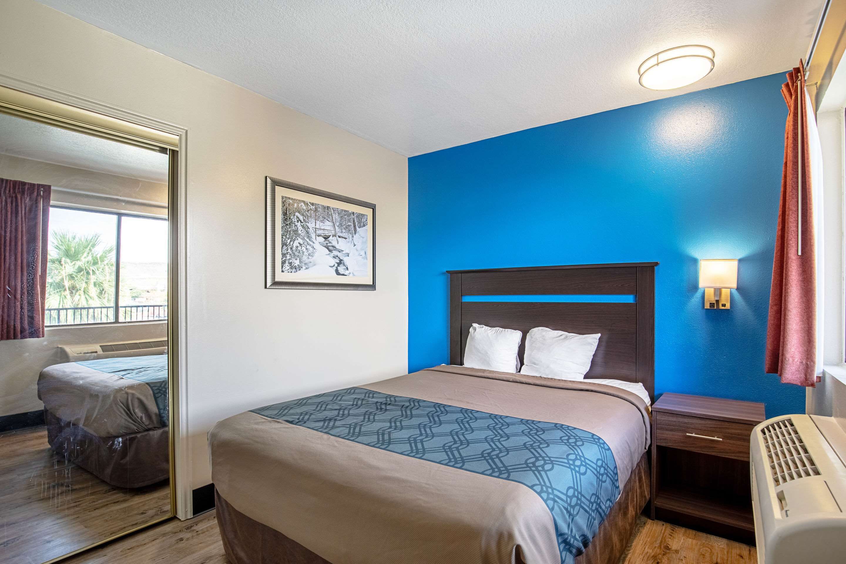 Una habitación de hotel con cama, mesita de noche, lámpara de pared y ventana con cortinas.