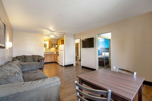 A living room with a gray sectional sofa, a wooden dining table with four chairs, and a kitchen in the background.