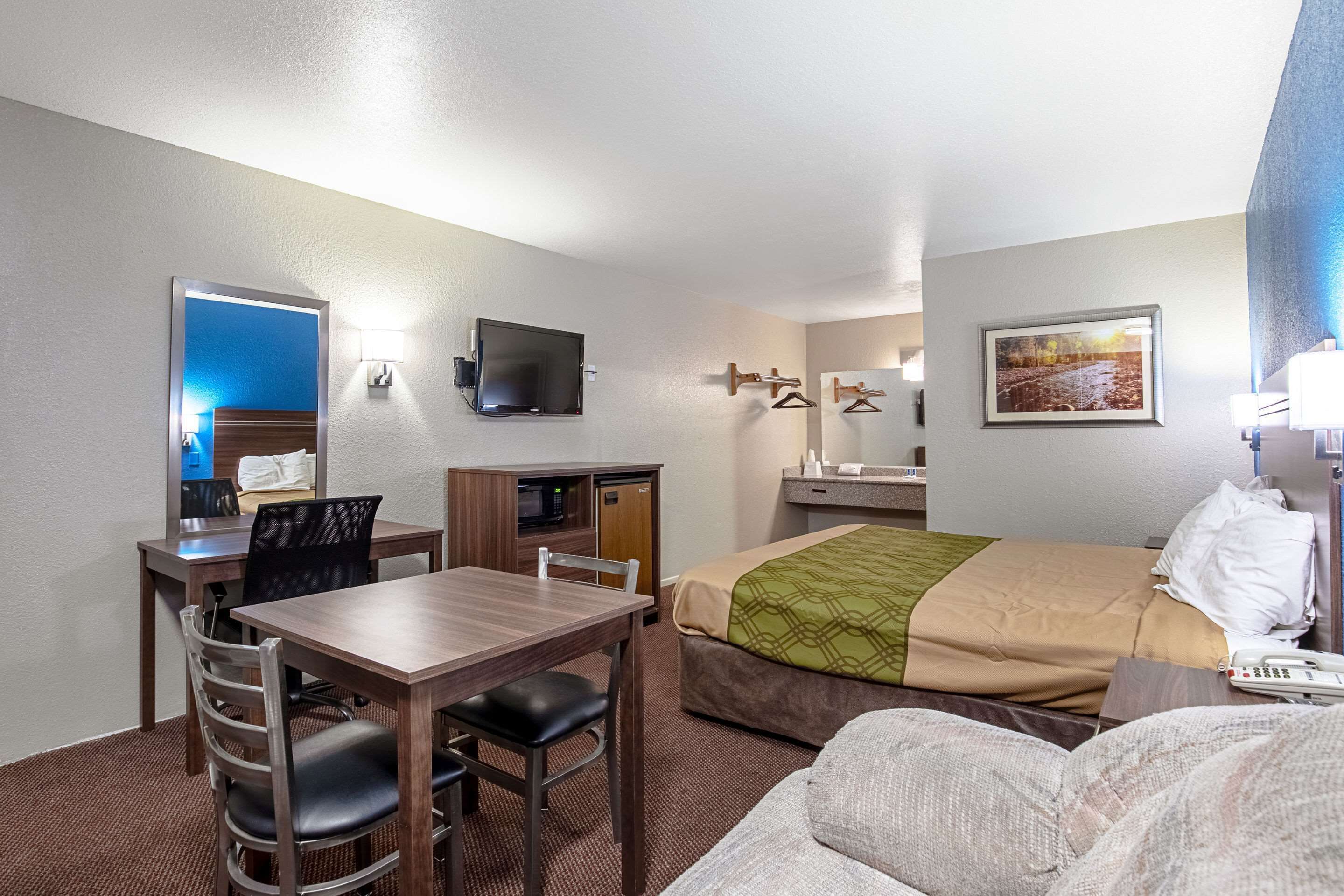 Quarto de hotel com cama, escrivaninha, cadeira, televisão, espelho, guarda-roupa e banheiro.