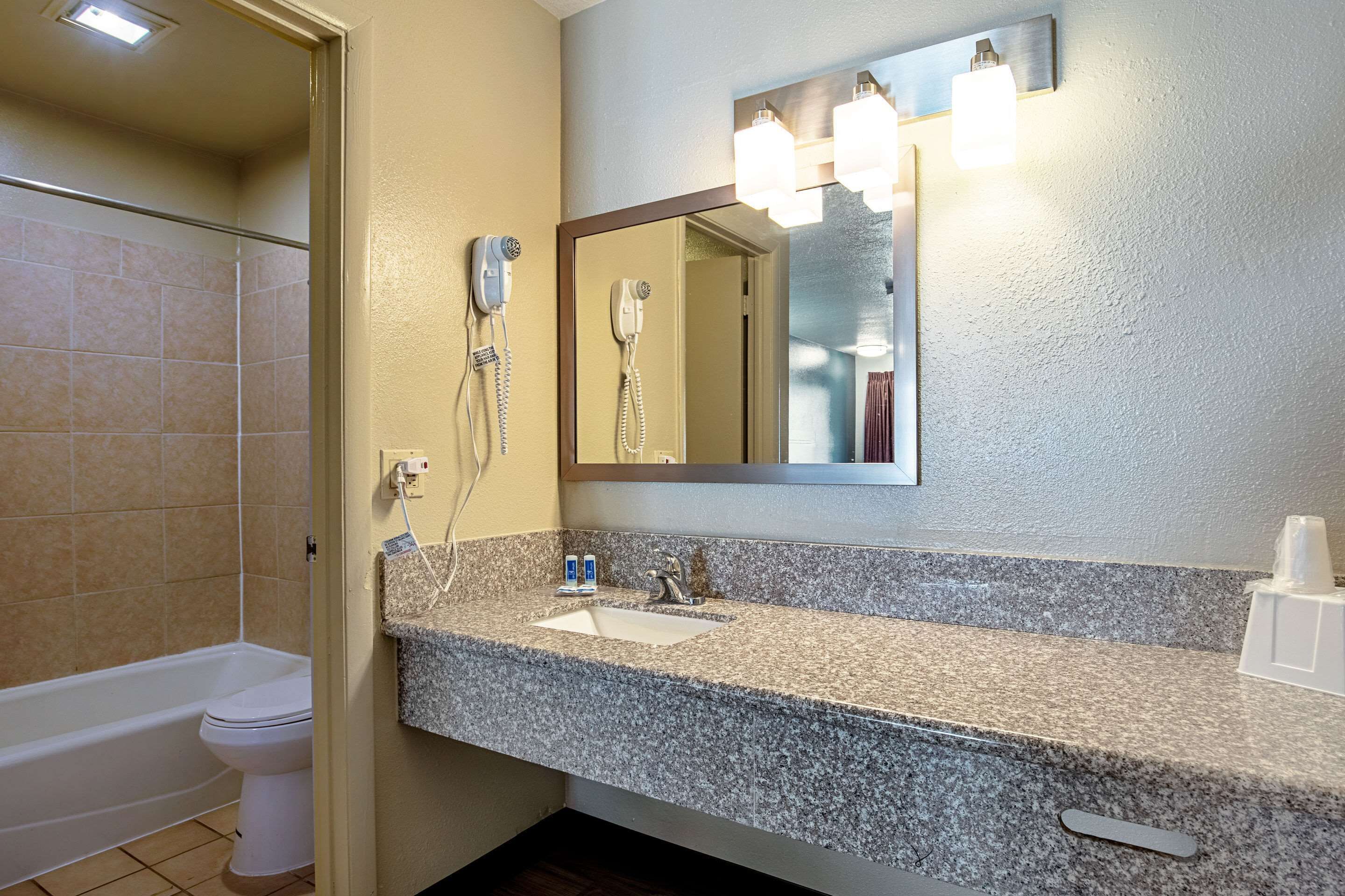 Un baño con encimera de granito, lavabo, espejo con iluminación, secador de pelo y ducha.