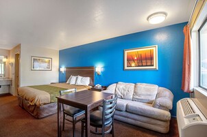Habitación de hotel con cama, sofá, mesa y escritorio. Se ve una ventana con cortinas, un reloj de pared y un marco de foto en la pared.